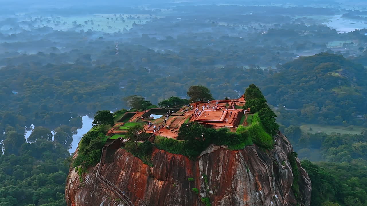 Multiple tourists walk by the ruins of the ancient stone fortress on the top of the picturesque mountain. Sigiriya palace, Sri Lanka, South Asia. Top view. Hazy scenery at backdrop.