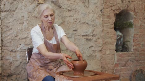 elderly female potter in an apron makes a pot in a pottery workshop