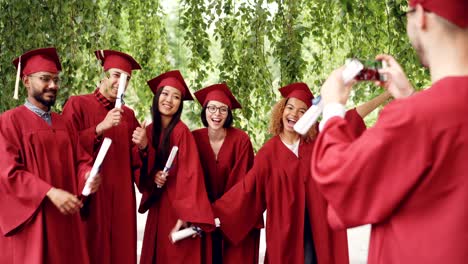 cheerful guy is using smartphone to take pictures of his fellow students graduates holding diplomas, posing and laughing. happiness, technology and education concept.