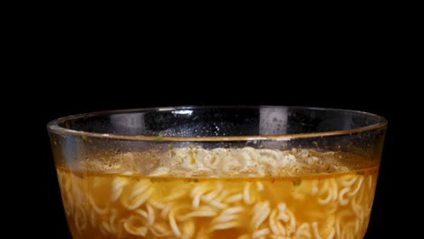 hand uses wooden chopsticks to lift cooked instant noodles from a clear glass bowl filled with broth, against a black background with even lighting