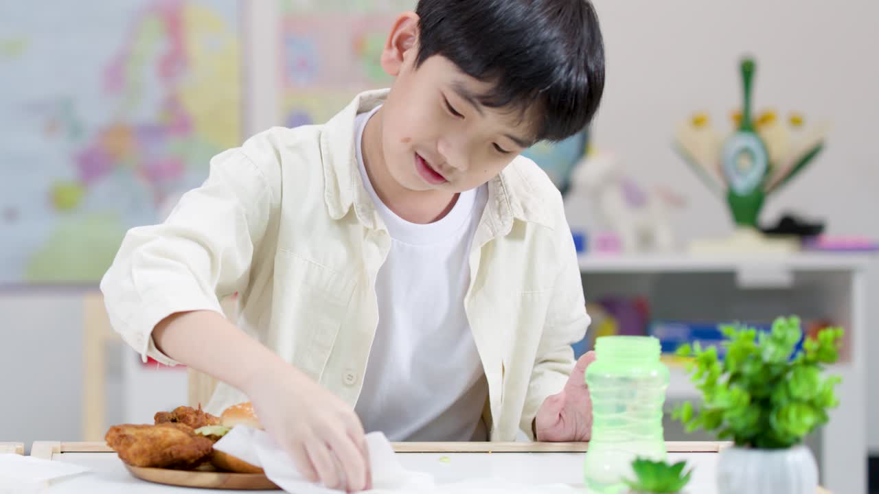 Young boy wipes classroom lunch table before eating fried chicken burger, bright static shot