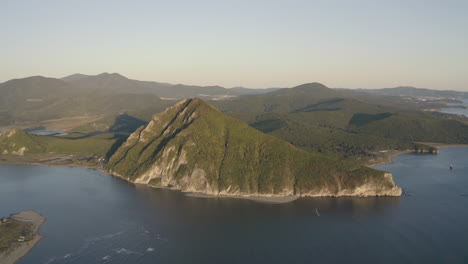 a landscape view of a pyramid-shaped mountain located at a river estuary flowing into the sea, with green vegetation on its sides, mountain ridge in the background, on the sunset