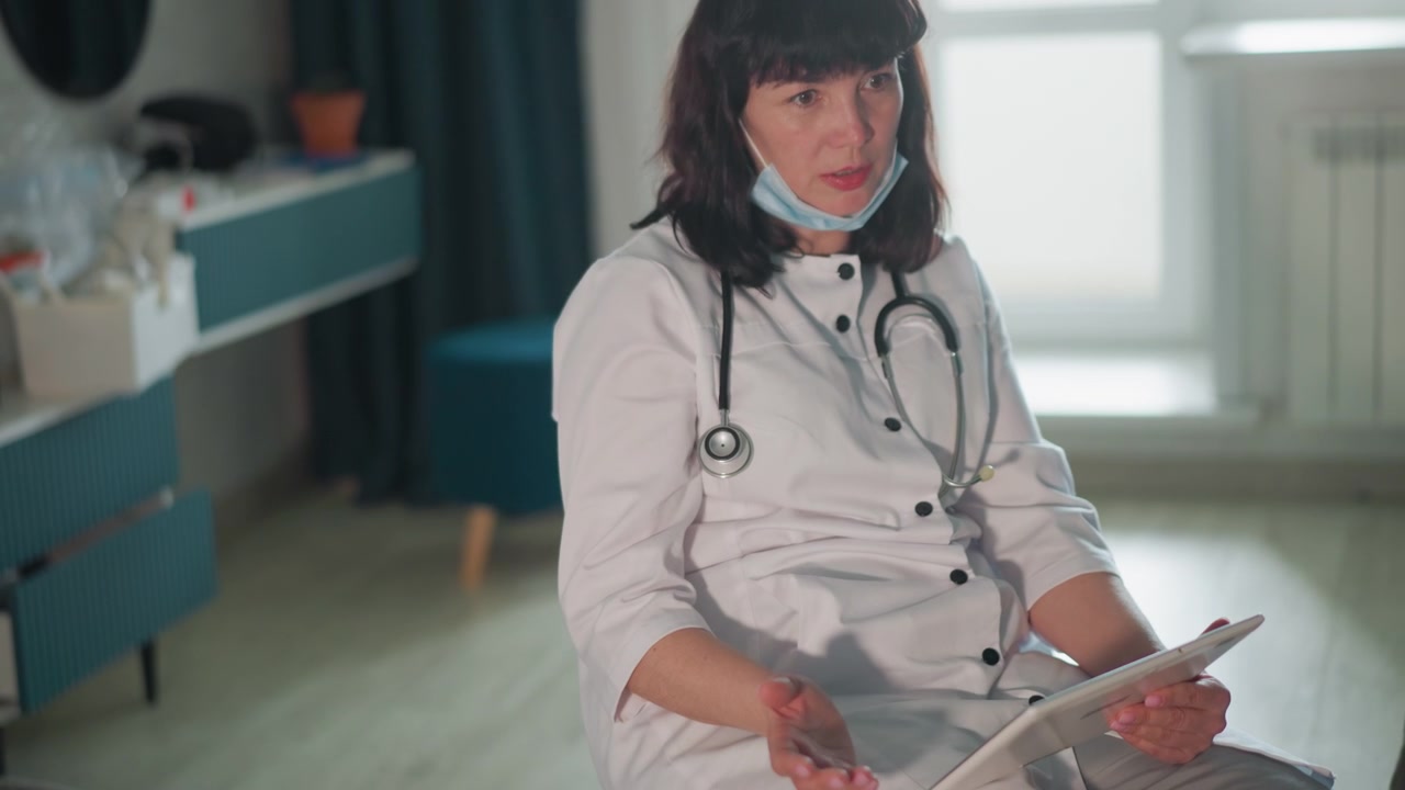Dottoressa bianca che effettua una visita domiciliare con un paziente sul divano, stetoscopio al collo, appunti su tablet visibili, valutazione tranquilla tramite conversazione, ascolto attento e rassicurazione professionale.