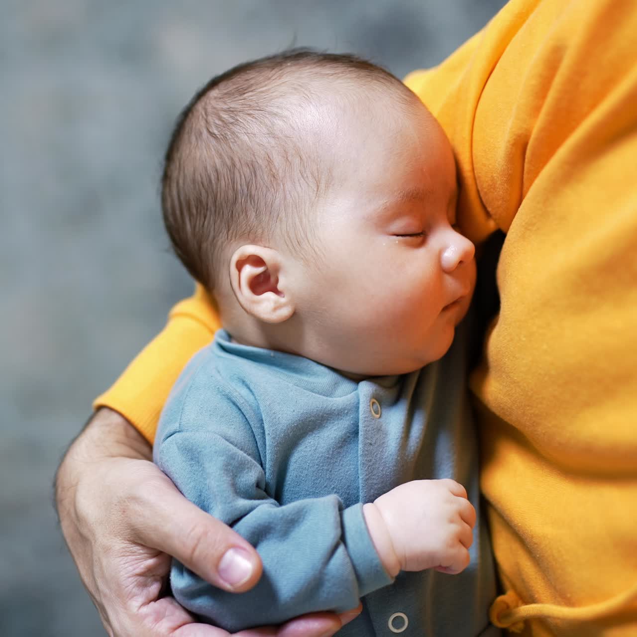Adorable baby boy in blue clothes resting peacefully in his parent's arms. Male in yellow sweater holding a sleeping baby. Close up