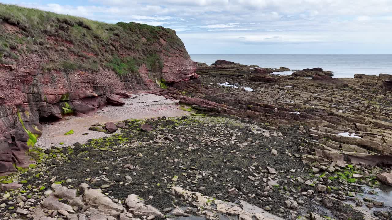 Camera slowly pans over red sandstone cliffs, rocky tidal shore, and seaweed at low tide
