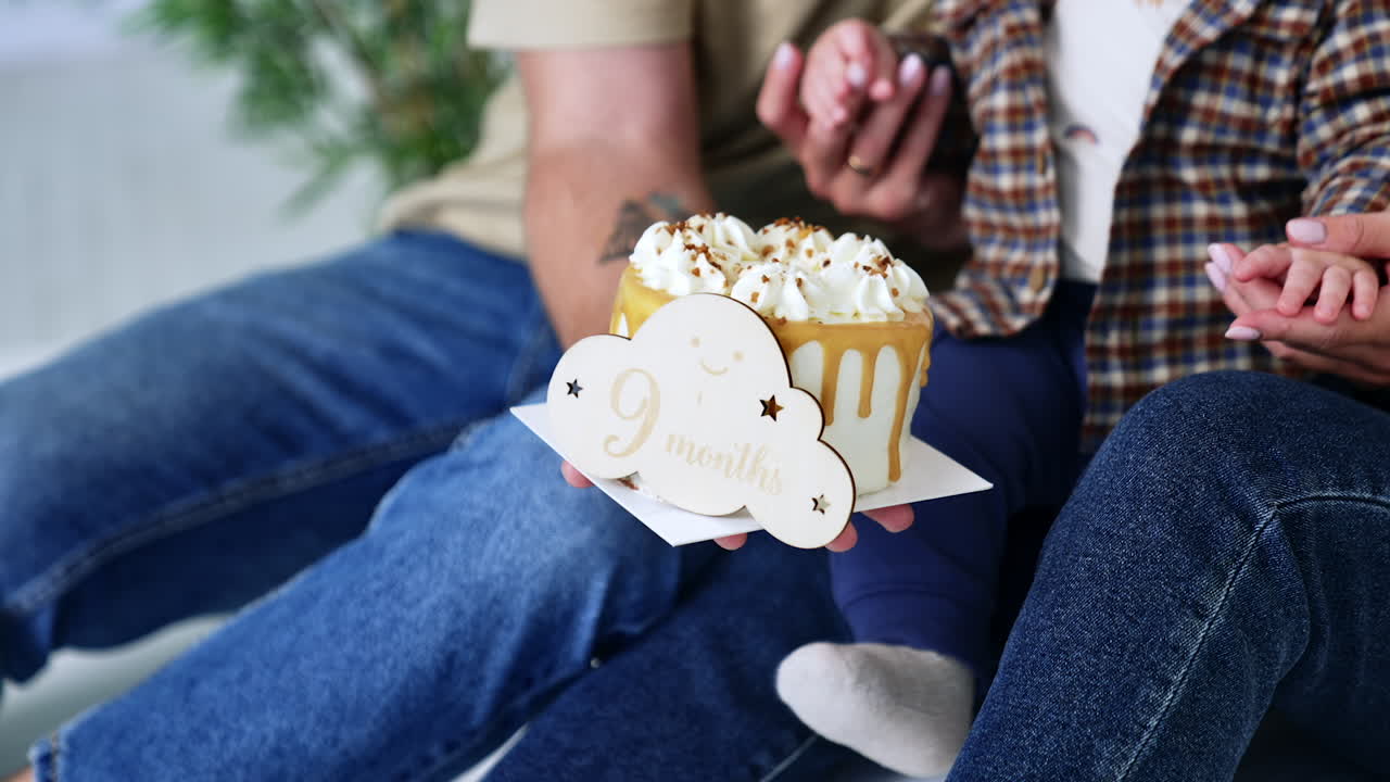 A wooden plate with 9 months inscription stands at the cake. Unrecognized family of three celebrates child's ninth month anniversary. Close up.