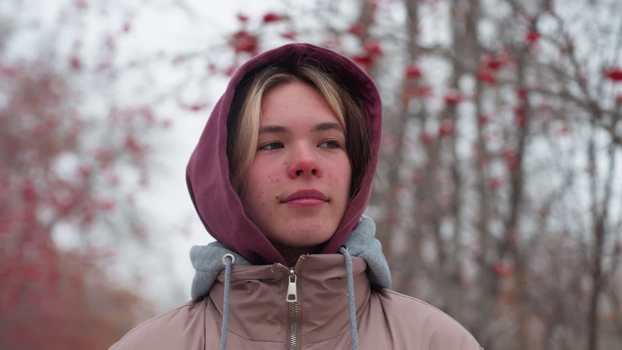 primer plano de una mujer con una chaqueta de invierno con capucha paseando con una cálida sonrisa, árboles cubiertos de nieve con bayas rojas crean una atmósfera de invierno pacífica y serena en el fondo borroso