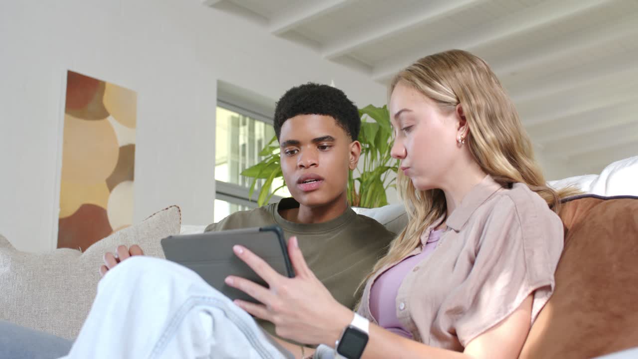 Diverse couple leaning on couch, swiping tablet, man pointing, woman showing smartphone, sharing