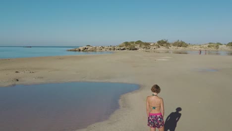 Vista-Aérea-De-Drones-De-Una-Mujer-Parada-En-El-Reflejo-De-La-Laguna-De-La-Playa-De-Elafonissi