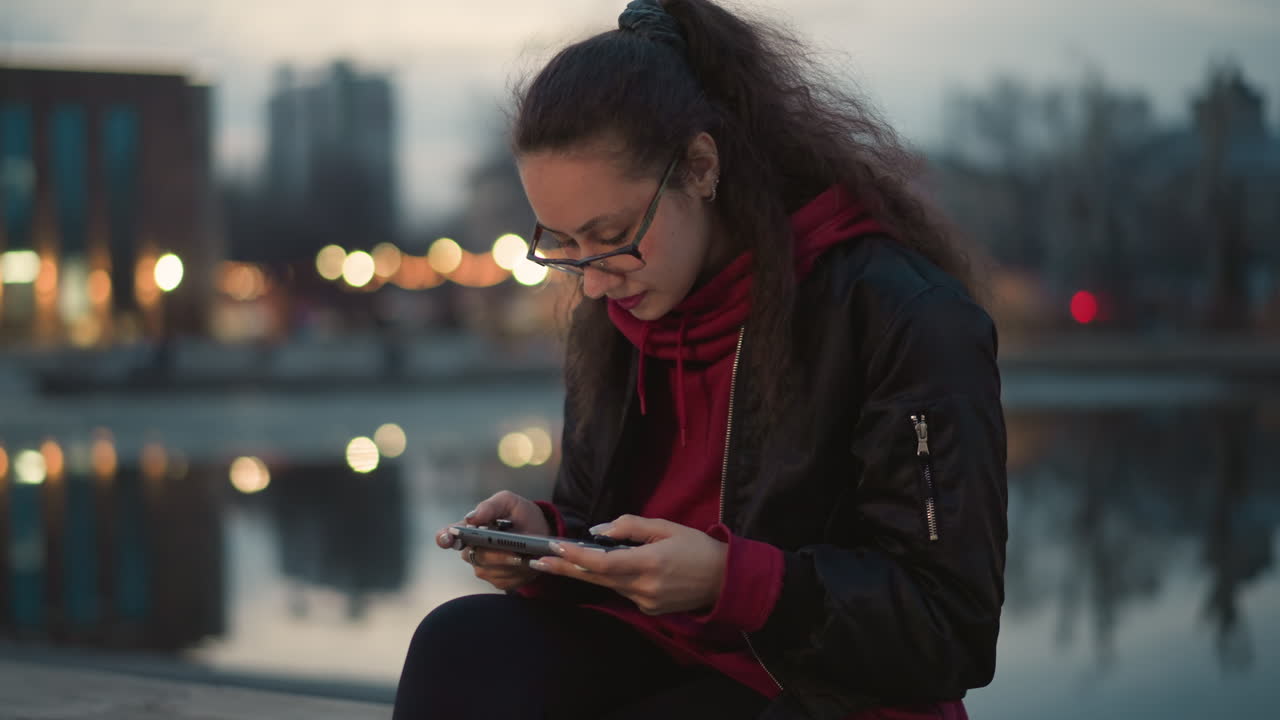 Mulher interagindo com o celular à beira-mar, mulher serena interagindo com um jogo perto da água à noite, mulher asiática sorri tranquilamente enquanto toca seu dispositivo móvel perto dos reflexos da orla