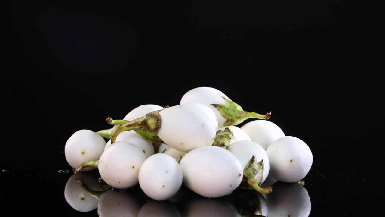 A cluster of Thai eggplants rotates on a glossy black surface under bright lighting, showcasing their smooth texture