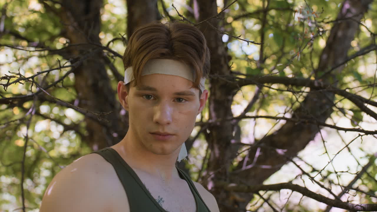 front view of soldier facing camera in sunlit forest expression lost and alert under leafy canopy branches framing profile vivid green background creating mood of solitude and cautious exploration