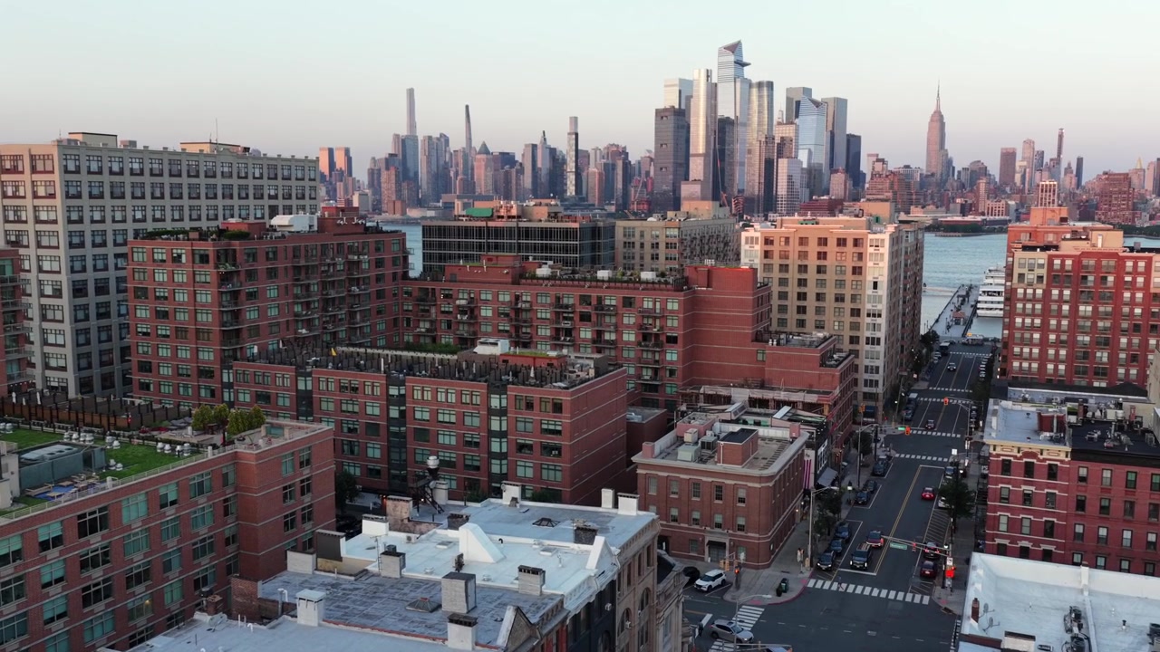 Røde murstenslejlighedsbygninger med taghaver fylder havnefronten i Hoboken, New Jersey, Hudson River og den fjerne Empire State Building gløder varmt under en gylden solnedgang. Droneflyvning nærmer sig.