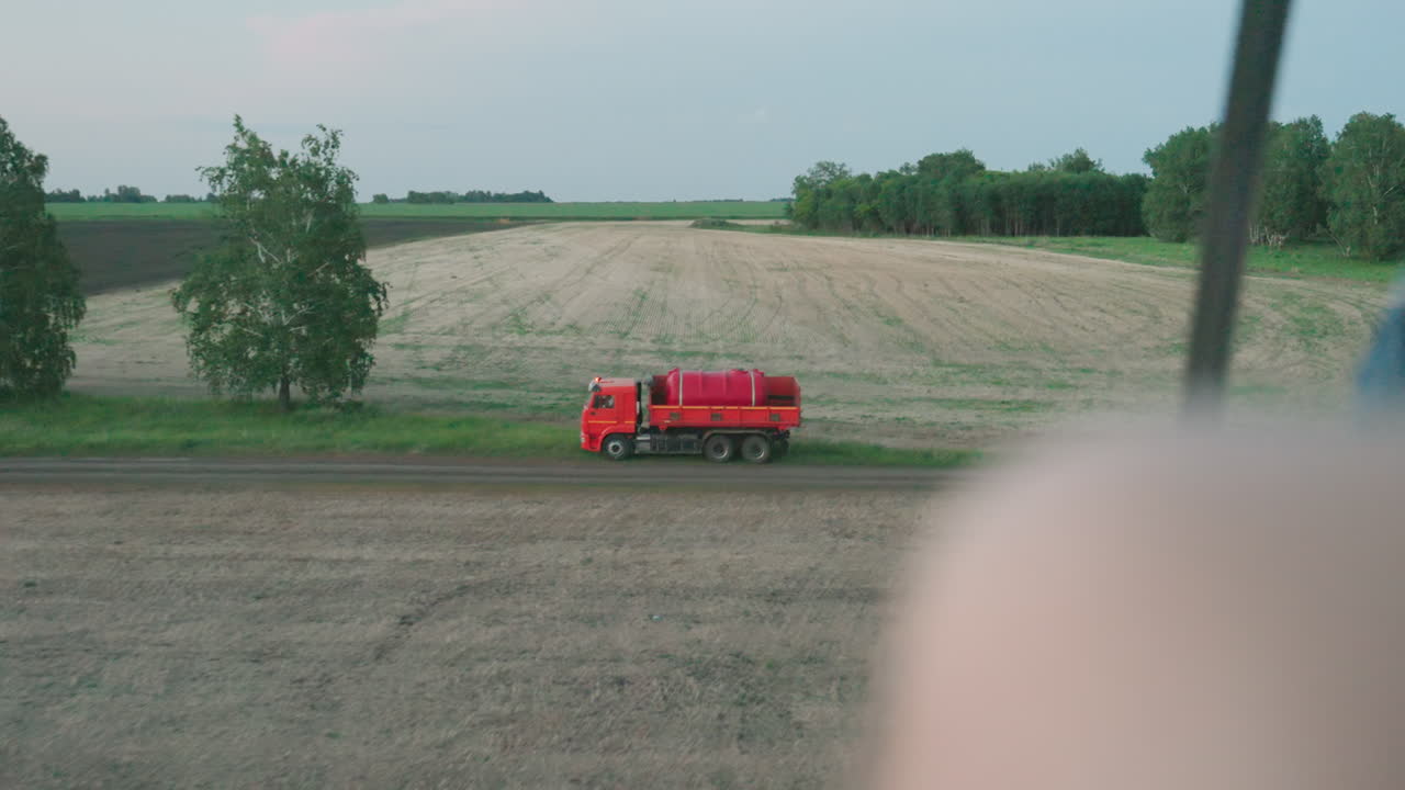 overhead drone view shows red tanker truck driving along dirt road beside harvested and green fields under soft evening sky, showcasing agricultural transport motion and contrast between vehicle