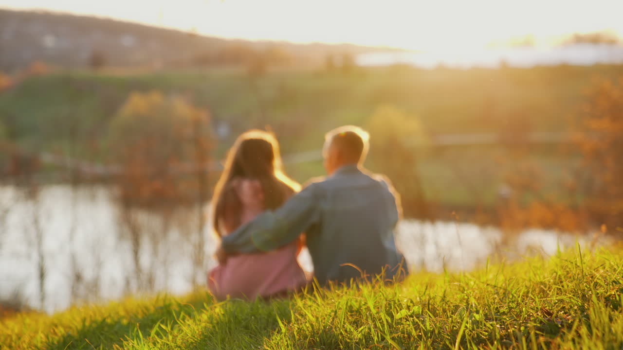 Romantic couple rests in nature. Young people boy and girl sitting together on grass and looking at sunset. Couple in love on date outdoors.