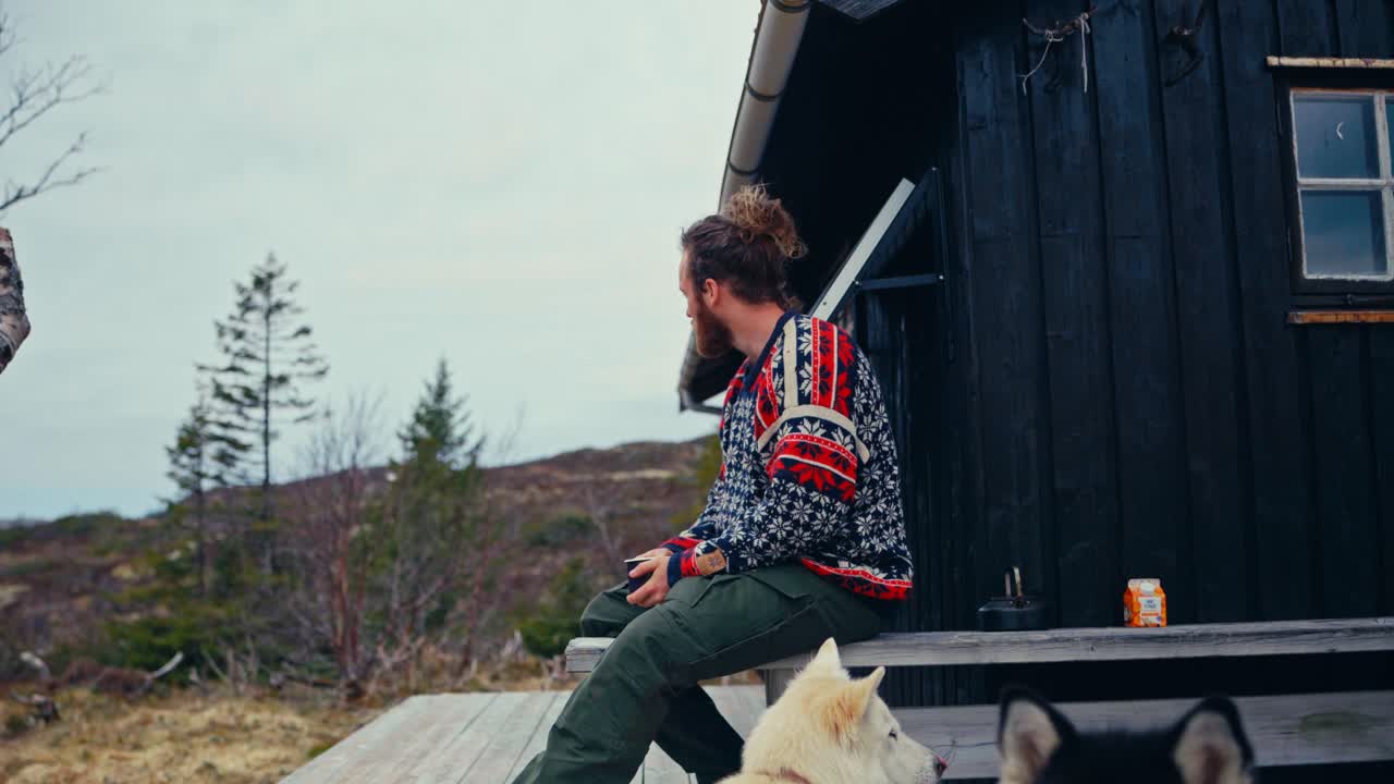 Man With A Beard Drinking Coffee Outside The Cabin With Alaskan Malamute Dogs. Static Shot