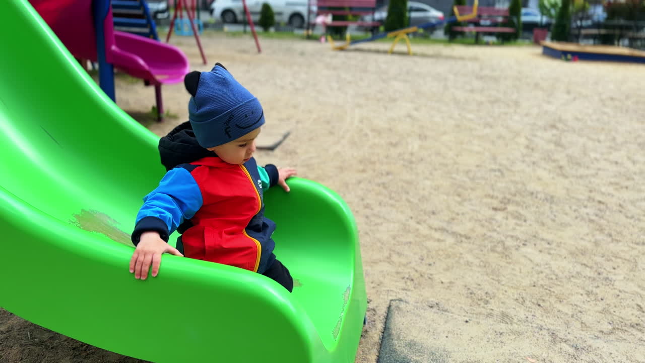 Cute boy sits on the green slide on the playground outdoors. Toddler stands on his feet and runs away.