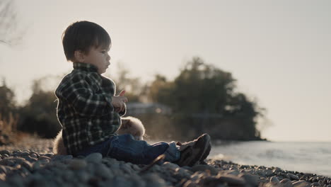 a little multi-ethnic boy sits on the shore of the lake next to the puppy and throws stones into the water.