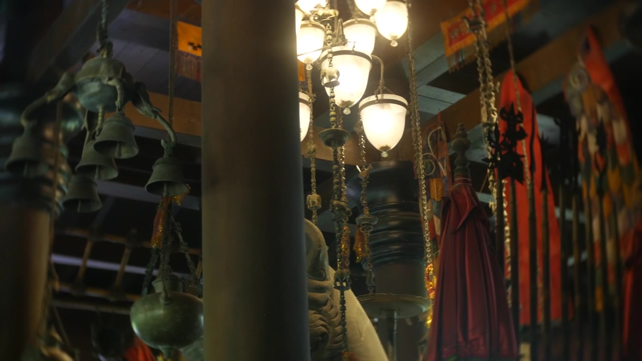 Bells hanging from a wooden structure inside a temple, small metal chimes suspended by chains around a central pillar. Warm interior lighting glowing against wood beams.