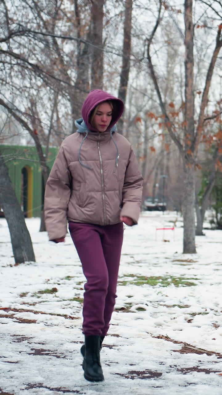 Young woman in winter outfit warms up in cold outdoor park, walking on snowy ground, dressed in hoodie, boots, and gloves, she moves through scenic park surrounded by leafless trees