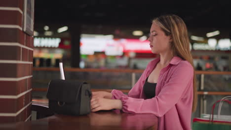 white lady in pink dress sits in a mall with her bag on a wooden table, opening her laptop to work, the background is blurred with people moving around, with a bokeh light effect