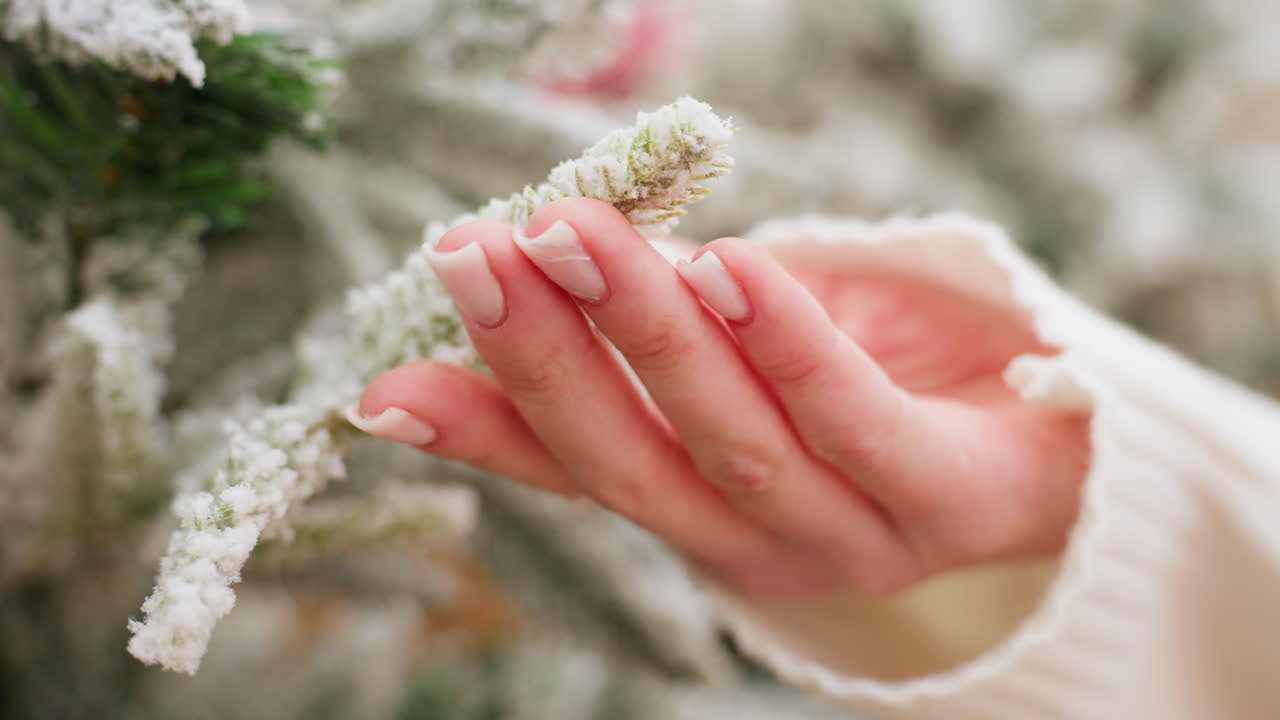 Close-up hand view of young lady in white sleeve gently touching snow-like Christmas tree in decorative store, with soft bokeh lights in background