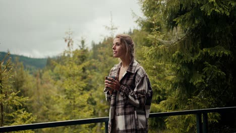 A-happy-blonde-girl-stands-with-a-cup-of-fragrant-hot-coffee-on-the-balcony-of-a-country-house-and-looks-at-the-mountain-coniferous-forest