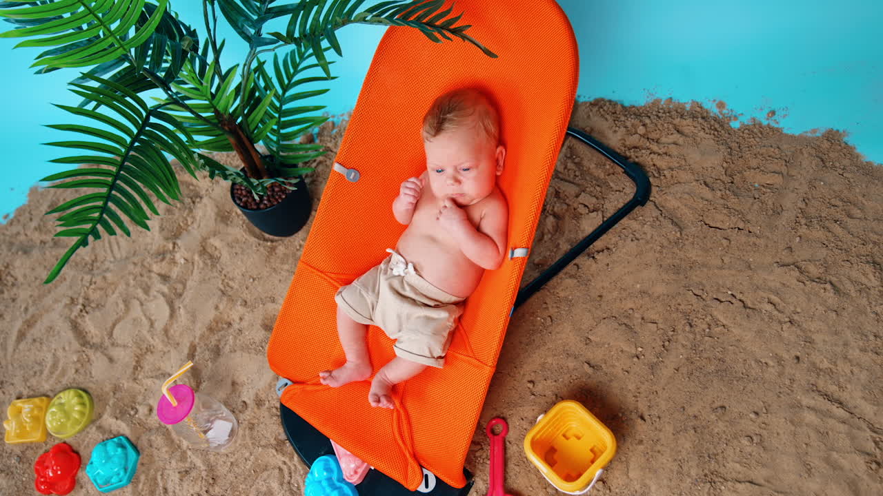 Calm baby boy having rest in the orange chair. Sand, plant in the pot and toys around. Top view.