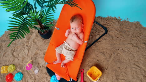 calm baby boy having rest in the orange chair. sand, plant in the pot and toys around. top view.