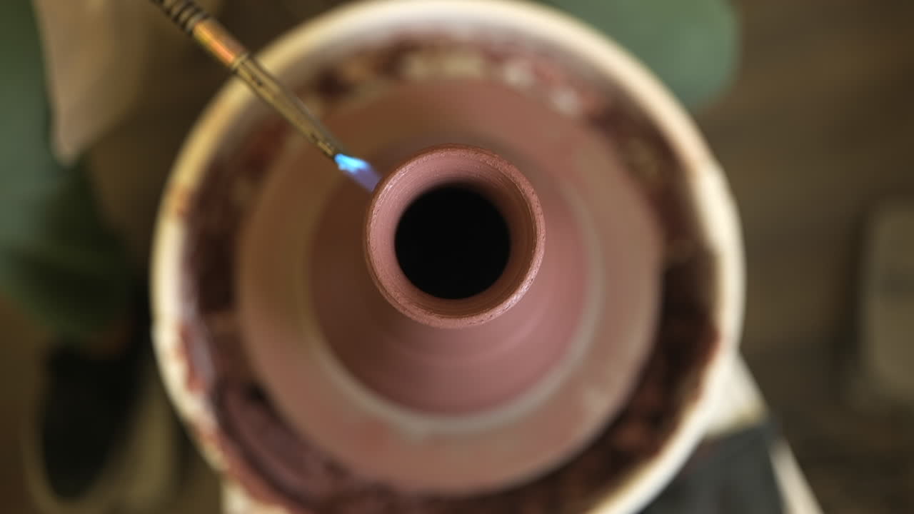 Shallow focus looking down over clay vase rotating on pottery wheel being glazed with propane torch