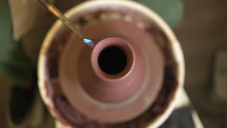 Shallow-focus-looking-down-over-clay-vase-rotating-on-pottery-wheel-being-glazed-with-propane-torch