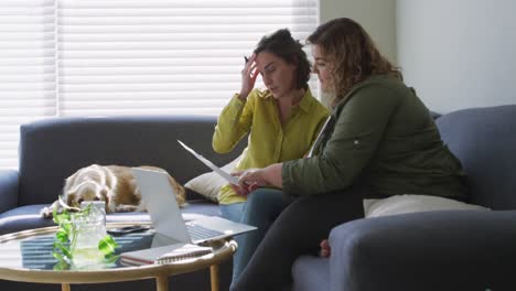 Caucasian-lesbian-couple-using-laptop-and-sitting-on-couch-with-dog