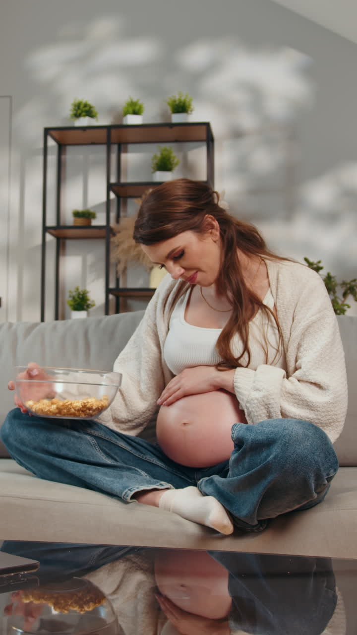 Pregnant woman sitting on couch eating popcorn from bowl relaxing at home