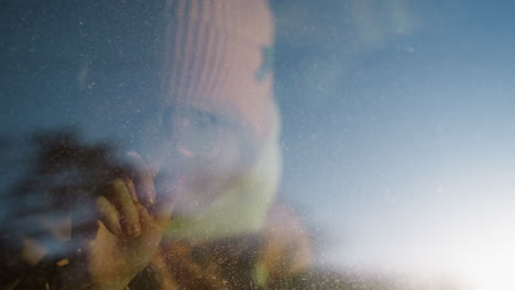 a young girl in a pink knit hat and scarf drawing with her finger on a foggy car window. her face is close to the glass, capturing a moment of creative play and curiosity on a cold, frosty day