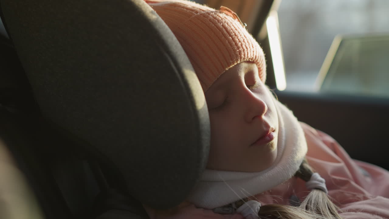 핑크색 뜨개질 모자와 스카프를 입은 어린 소녀가 차 좌석에서 평화롭게 잠을 자고 있습니다. 빛은 그녀의 조용한 얼굴을 부드럽게 비추고 있습니다.