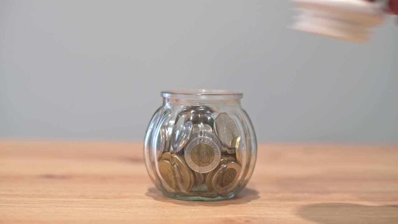 A close-up of a hand dropping a coin into a glass jar filled with coins, symbolizing saving money, financial growth, or budgeting.