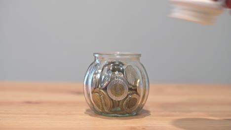 A close-up of a hand dropping a coin into a glass jar filled with coins, symbolizing saving money, financial growth, or budgeting.