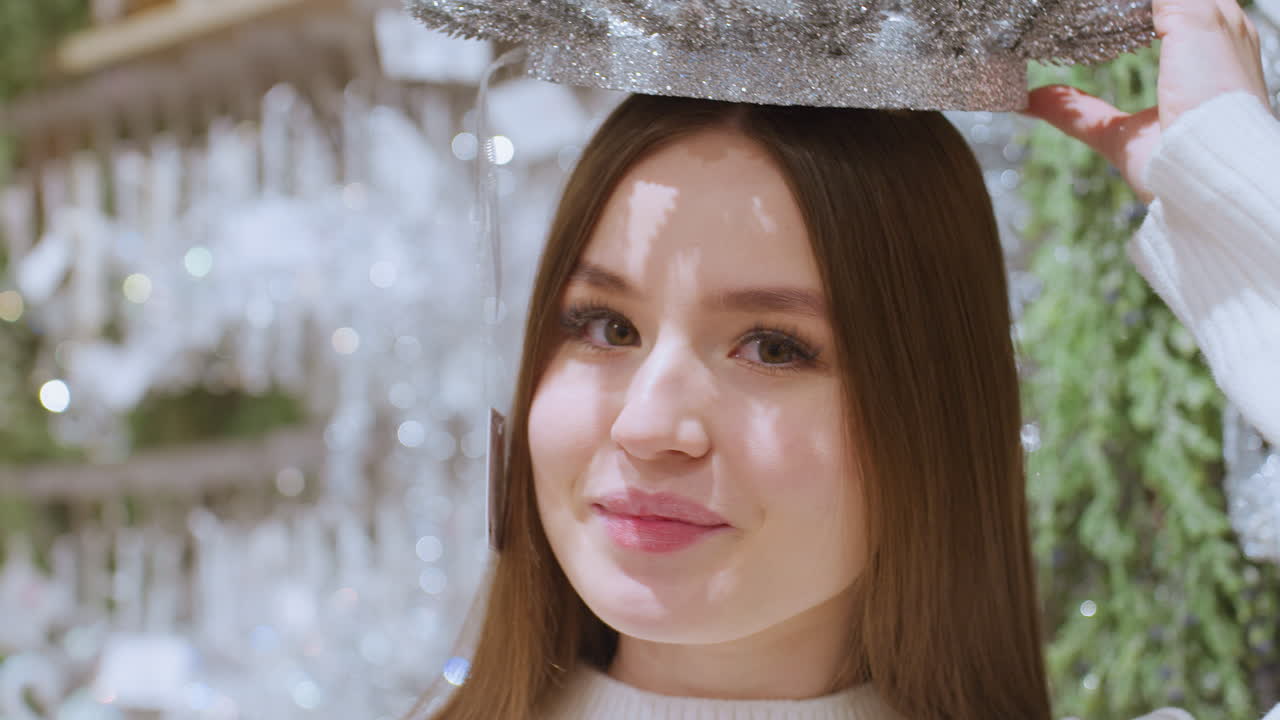 Young woman playfully places sparkling Christmas decoration on her head, surrounded by festive decor in store, enjoying holiday shopping in a beautifully decorated retail environment