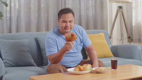 a fat asian man looking at fried chicken in his hand then smiling and showing thumbs up gesture to camera while eating fast food on a sofa in the living room at home