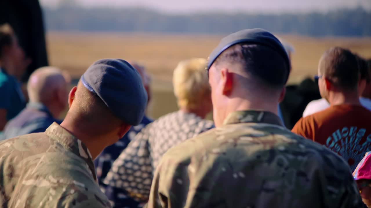 Two soldiers in camouflage uniforms with berets observe a crowd at the Airborne Ede event. The background includes civilians and a relaxed atmosphere on a bright, sunny day.