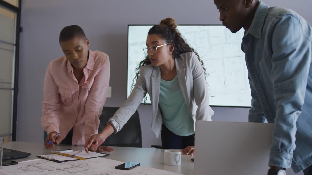 Three diverse male and female creative colleagues in discussion standing at table in meeting room