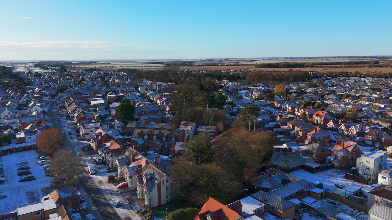 Toma aérea captura un pueblo cubierto de nieve con casas de tejados rojos. Cielo azul claro enmarca el río y los campos distantes. Árboles otoñales con hojas doradas contrastan con la nieve