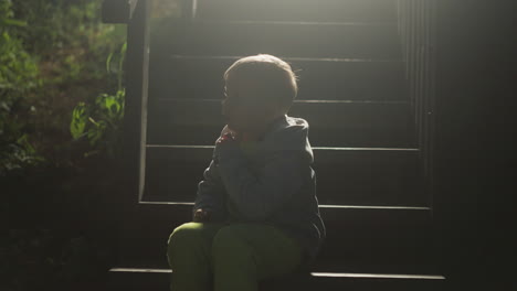 little child sitting on stairs silhouette in evening. boy enjoys calm night resting on steps at country house. kid spends time on pouch at backlight