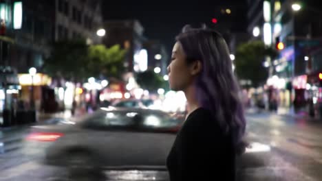 a young woman with purple hair stands in the middle of an empty city street at night, illuminated by colorful lights from nearby storefronts. urban atmosphere feels calm and reflective.