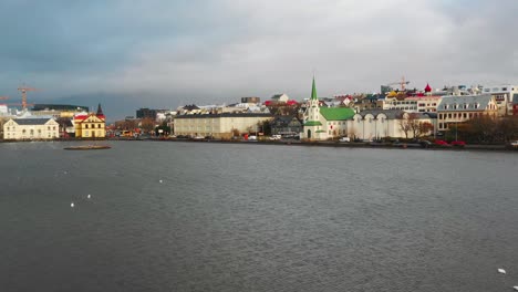 Aerial-View-of-Reykjavik,-Iceland,-Lake-Tjornin-Lakefront,-Frikrikjan-Lutheran-Church-Landmark-and-Cityscape-on-Cloudy-Summer-Day,-Drone-Shot