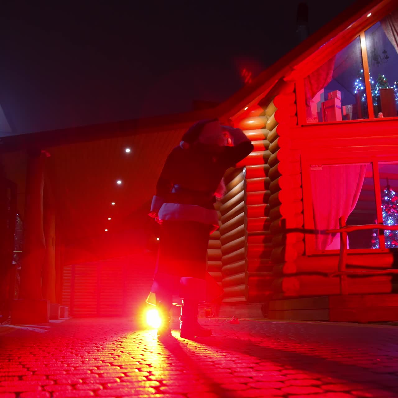 Santa Claus with bag full of presents. Santa Clause walking to the decorated houses with lights and carrying Christmas gifts. Red lighting. Back view