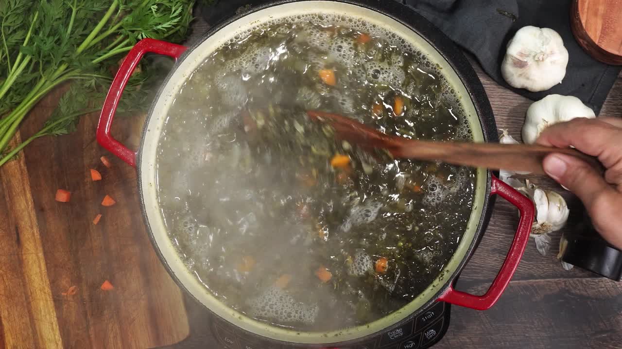Cooking Italian Wedding Soup and adding in the baked tiny meatballs into the red stock pot. Little meatballs being added into a simmering pot. Using a wooden spoon to mix the soup while its simmering.