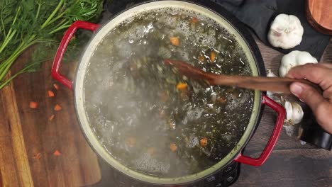 cooking italian wedding soup and adding in the baked tiny meatballs into the red stock pot. little meatballs being added into a simmering pot. using a wooden spoon to mix the soup while its simmering.