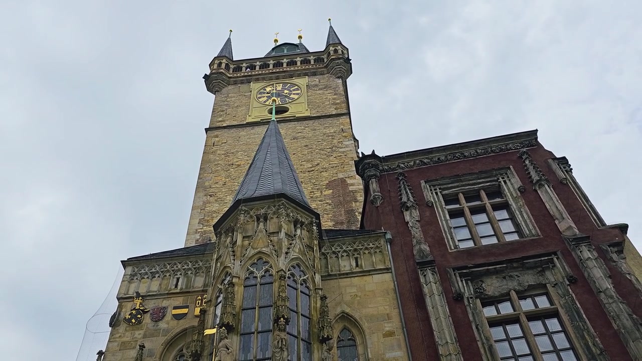 Historic Prague Old Town Hall tower filmed from below, featuring the famous astronomical clock, Gothic spires, detailed stone facade, red building, and overcast Czech city sky.