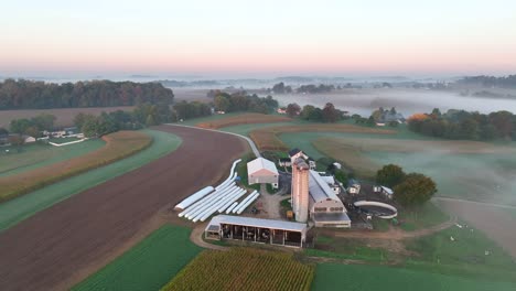 aerial dawn view of a farm with silos, multiple barns, patchwork fields, a house, and fog